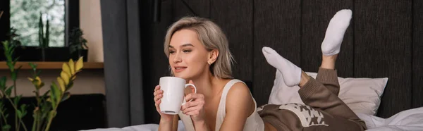 Панорамный снимок привлекательной улыбающейся женщины, держащей чашку чая во время отдыха на кровати — стоковое фото