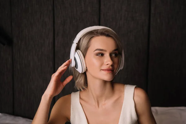 Mujer atractiva tocando auriculares inalámbricos mientras mira hacia otro lado - foto de stock