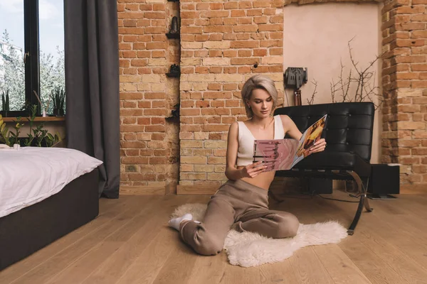 Belle jeune femme assise sur un tapis moelleux dans la chambre et le magazine de lecture — Photo de stock