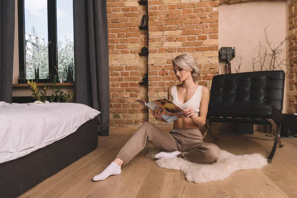 Hermosa mujer joven sentada en la alfombra lanuda en el dormitorio y el diario Reding - foto de stock