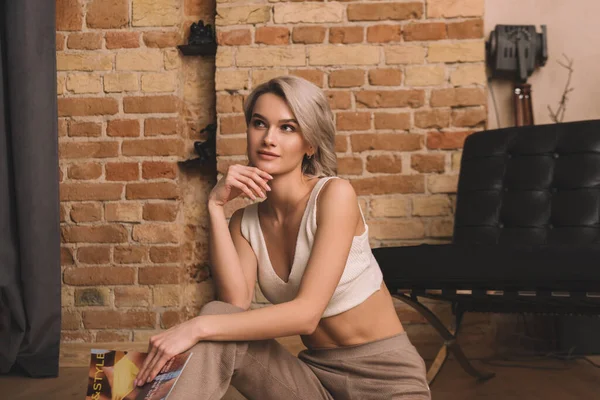 Привлекательная молодая женщина сидит на полу, держит журнал и смотрит вдаль на дом — стоковое фото