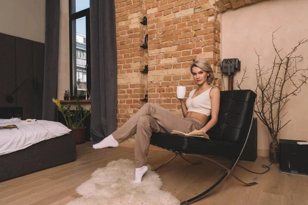 Attractive woman holding cup of tea and looking at camera while sitting in armchair near book at home — Stock Photo