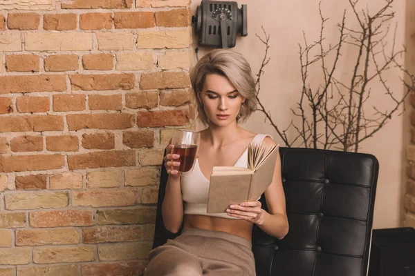 Femme concentrée tenant un verre de vin rouge tout en lisant le livre à la maison — Photo de stock