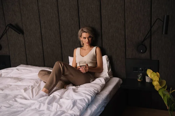 Bella, donna fiduciosa in possesso di un bicchiere di vino rosso mentre seduto sul letto e guardando altrove — Foto stock