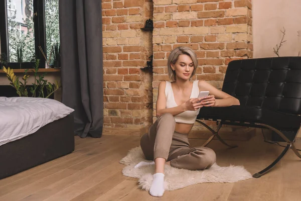 Schöne junge Frau plaudert auf Smartphone, während sie auf flauschigem Teppich im Schlafzimmer sitzt — Stockfoto