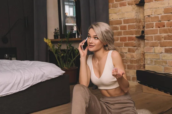 Sorridente, donna attraente che parla sullo smartphone mentre siede sul pavimento in camera da letto — Foto stock