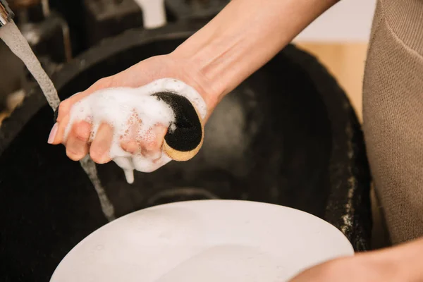 Cropped view of woman holding soapy sponge near white plate — Stock Photo