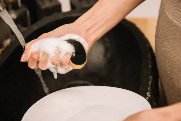 Vista ritagliata della donna che tiene spugna sapone vicino piatto bianco — Foto stock
