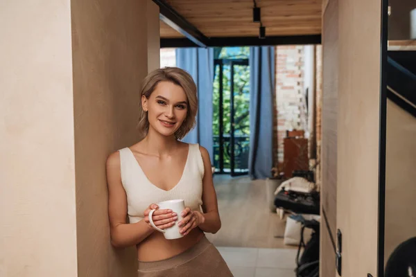 Счастливая, привлекательная женщина, стоящая у стены дома и улыбающаяся на камеру, держа чашку чая — стоковое фото