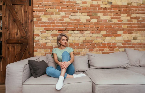 Приваблива, вдумлива жінка, сидить на дивані біля цегляної стіни і дивиться в сторону — стокове фото