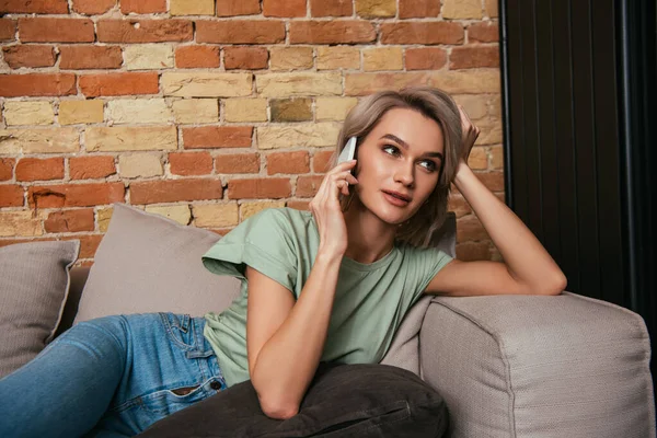 Bella donna guardando lontano mentre seduto sul divano e parlando su smartphone — Foto stock