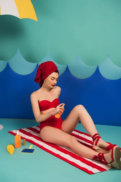Atractiva mujer en toalla roja y traje de baño aplicando protector solar cerca de las ondas de corte de papel - foto de stock