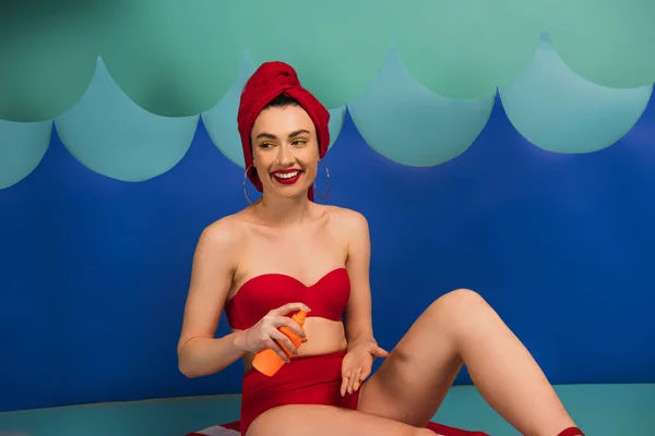 Femme heureuse en serviette rouge et maillot de bain appliquant crème solaire près des vagues de coupe de papier — Photo de stock
