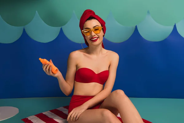 Happy woman in red swimsuit and sunglasses holding sunscreen near paper cut waves — Stock Photo