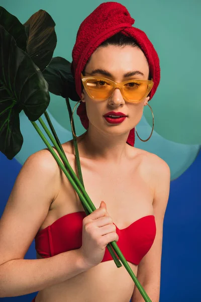 Attractive girl in red towel swimsuit and sunglasses holding tropical palm leaves on blue and turquoise — Stock Photo