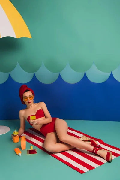 Attraente donna in costume da bagno rosso e occhiali da sole utilizzando smartphone vicino onde taglio carta — Foto stock