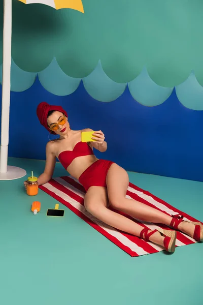 Привлекательная женщина в купальниках и солнцезащитных очках делает селфи рядом с волнами от резки бумаги — стоковое фото