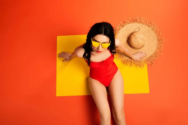 Top view of young woman in sunglasses and swimsuit sitting near straw hat on orange — Stock Photo