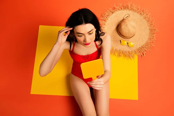Vista dall'alto della giovane donna in costume da bagno guardando lo specchio mentre seduta vicino al cappello di paglia e occhiali da sole su arancione — Foto stock