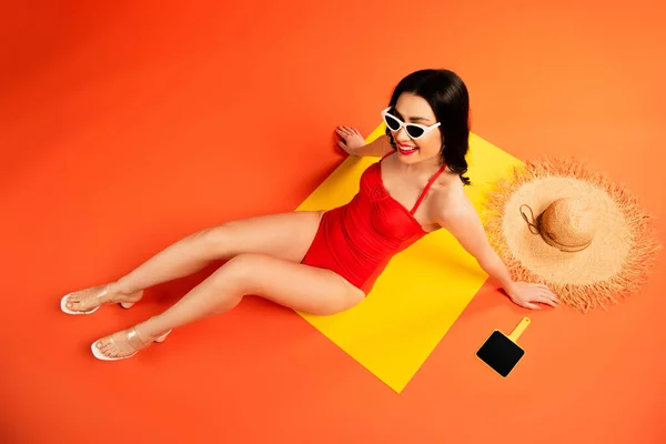 Top view of cheerful woman in sunglasses and swimsuit sitting near straw hat and mirror on orange — Stock Photo