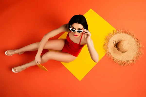 Vista superior de la mujer en traje de baño tocando gafas de sol y sentado cerca de sombrero de paja y espejo en naranja - foto de stock