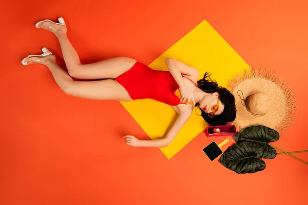 Vista dall'alto della donna in costume da bagno e occhiali da sole che applica la protezione solare vicino a cappello di paglia, foglie di palma e specchio su arancione — Foto stock