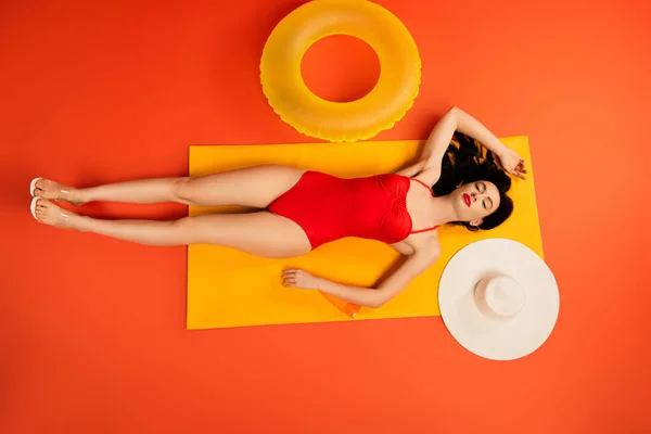 Вид сверху на девушку в купальнике лежащую рядом с бутылкой с солнцезащитным кремом, надувным кольцом, соломенной шляпой и зеркалом на оранжевом — стоковое фото
