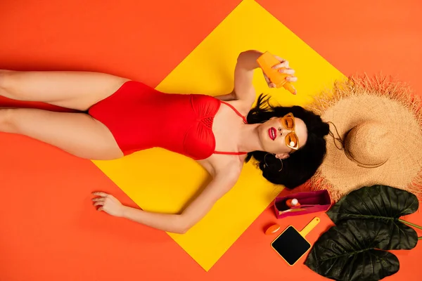 Vista superior de la chica en traje de baño y gafas de sol sosteniendo botella con protector solar cerca de sombrero de paja, hojas de palma y espejo en naranja - foto de stock
