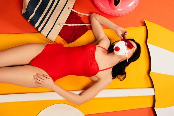 Vista dall'alto di bella donna in occhiali da sole, maschera protettiva e costume da bagno sdraiato vicino alla borsa e ombrello di carta su arancione — Foto stock