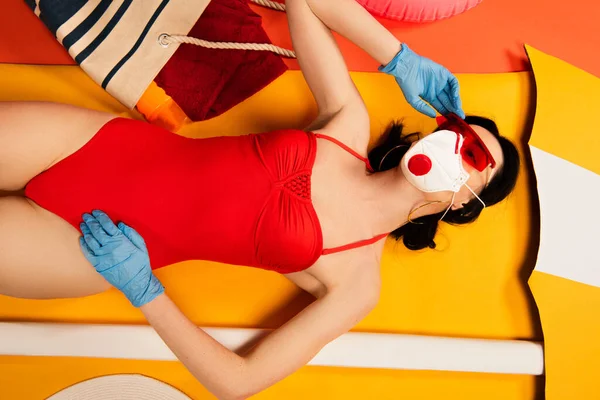 Top view of woman in sunglasses, medical mask, latex gloves and swimsuit lying near bag on yellow — Stock Photo