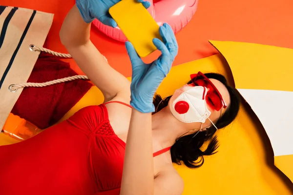 Donna in occhiali da sole, maschera medica, guanti in lattice e costume da bagno scattando selfie vicino alla borsa su arancione — Foto stock