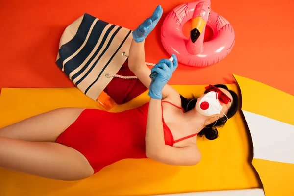 Frau mit Sonnenbrille, medizinischer Maske, Latexhandschuhen und Badeanzug trägt Händedesinfektionsmittel auf Orange auf — Stockfoto