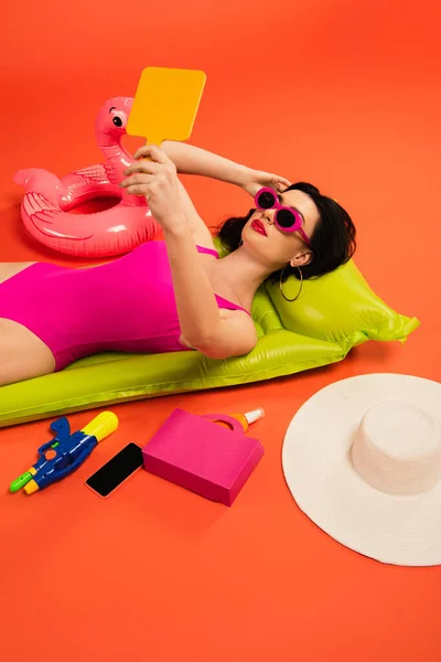 Woman in sunglasses and swimsuit lying on inflatable mattress and looking at mirror near inflatable ring and smartphone with blank screen on orange — Stock Photo