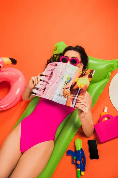 Mujer joven en gafas de sol y traje de baño acostado en el colchón inflable y cubriendo la cara con revista en naranja - foto de stock