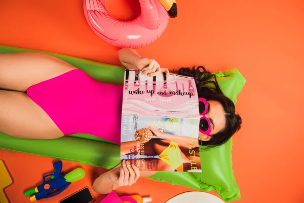 Vista superior de la mujer joven en gafas de sol y traje de baño acostado en el colchón inflable y cubriendo la cara con la revista en naranja - foto de stock