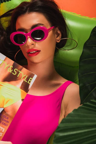 Vista dall'alto di attraente giovane donna in occhiali da sole e costume da bagno sdraiato su materasso gonfiabile con rivista vicino foglie di palma verde su arancione — Foto stock