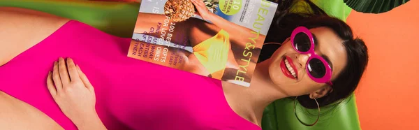 Panoramic crop of cheerful young woman in sunglasses and swimsuit lying on inflatable mattress with magazine near palm leaves on orange — Stock Photo