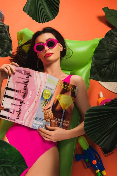 Top view of beautiful woman in sunglasses and swimsuit lying on inflatable mattress with magazine near palm leaves on orange — Stock Photo