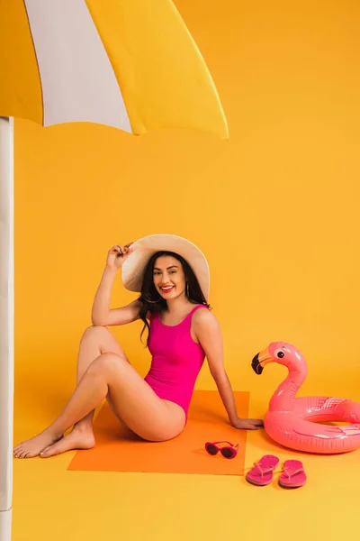 Happy woman in straw hat and swimsuit sitting near sunglasses, flip flops, inflatable ring and paper umbrella on yellow — Stock Photo