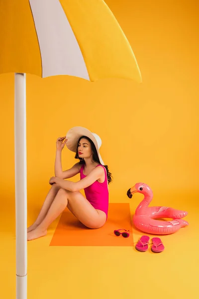 Attractive woman in straw hat and swimsuit sitting near sunglasses, flip flops, inflatable ring and paper umbrella on yellow — Stock Photo