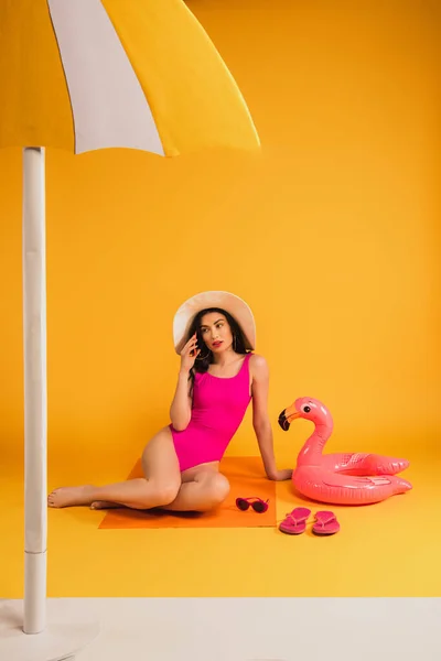 Young woman in straw hat and swimsuit sitting near sunglasses, flip flops, inflatable ring and talking on smartphone on yellow — Stock Photo