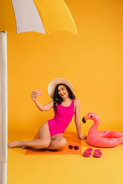 Happy woman in straw hat and swimsuit sitting near sunglasses, flip flops, and inflatable ring while taking selfie on yellow — Stock Photo