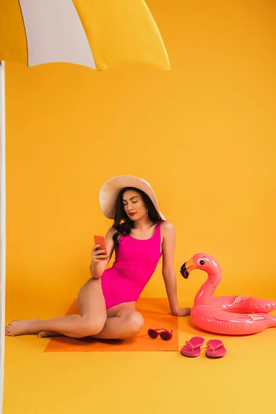 Attractive woman in straw hat and swimsuit sitting near sunglasses, flip flops and inflatable ring while using smartphone on yellow — Stock Photo