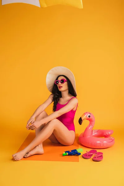 Молода жінка в солом'яному капелюсі, сонцезахисних окулярах і купальнику, що сидить біля надувного кільця, фліп-флоп і водяного пістолета на жовтому — стокове фото