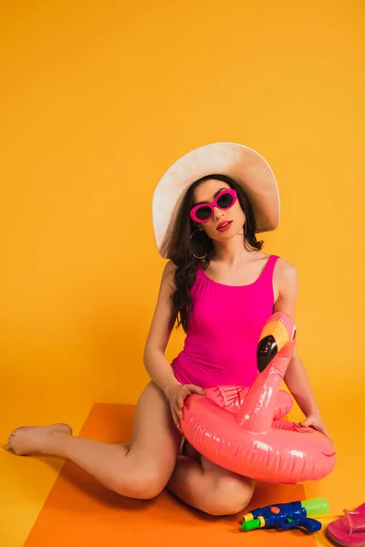 Woman in straw hat, sunglasses and swimsuit holding inflatable ring near water gun on yellow — Stock Photo