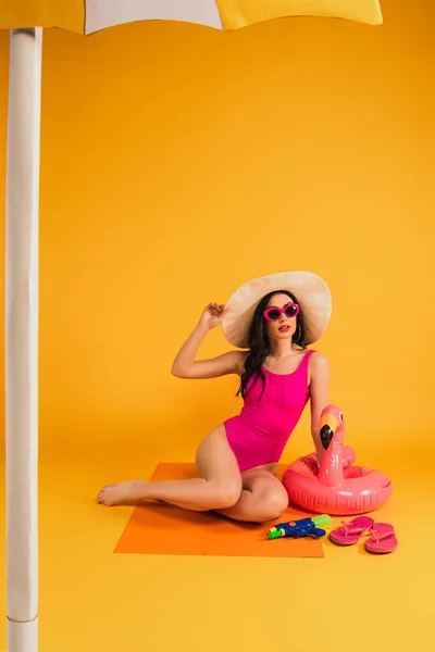 Mujer en sombrero de paja, gafas de sol y traje de baño tocando anillo inflable y chanclas cerca de pistola de agua en amarillo - foto de stock