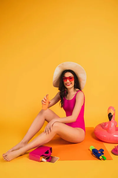 Happy woman in straw hat, sunglasses and bathing suit holding bottle with sunscreen near inflatable ring and water gun on yellow — Stock Photo