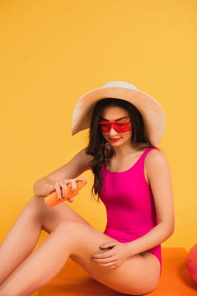 Donna con cappello di paglia, occhiali da sole e costume da bagno che applica la protezione solare sul giallo — Foto stock