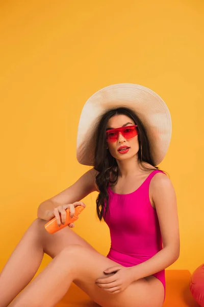 Жінка в солом'яному капелюсі, сонцезахисні окуляри та купальний костюм, що тримає пляшку з сонцезахисним кремом на жовтому — стокове фото