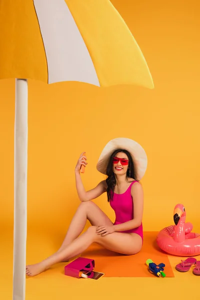 Mujer alegre en sombrero de paja, gafas de sol y traje de baño sosteniendo botella con protector solar cerca de anillo inflable y pistola de agua en amarillo - foto de stock